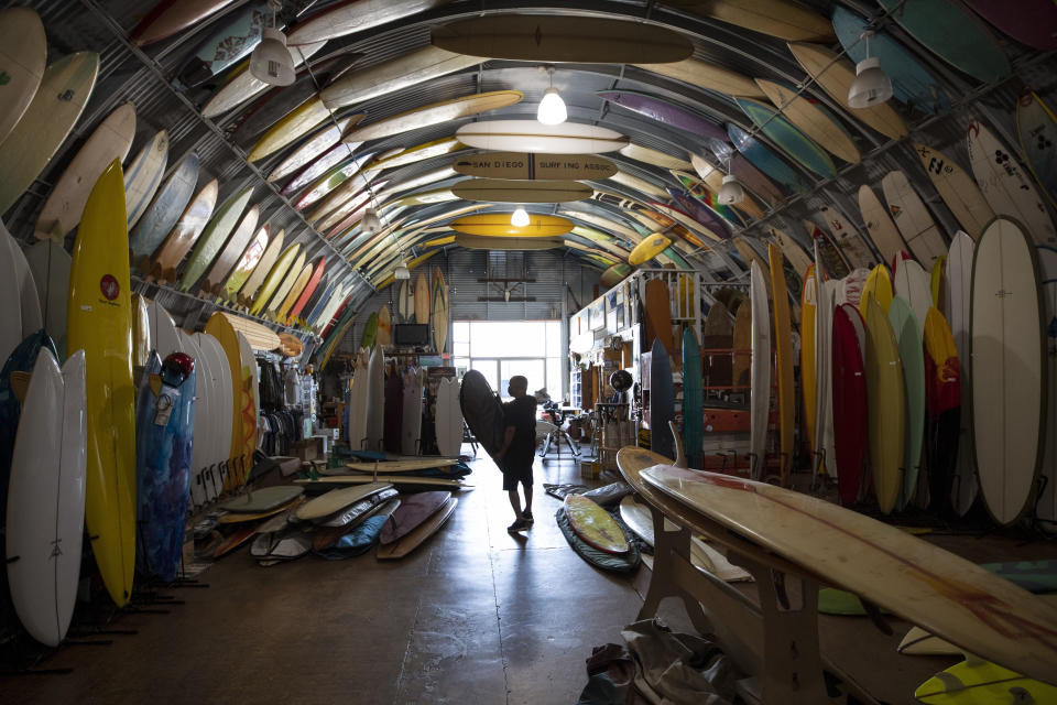 Surf shop worker Francois wears a mask as he carries a surfboard as Bird's Surf Shed opens Friday, May 8, 2020, in San Diego. California Gov. Gavin Newsom on Thursday issued the broadest loosening of his stay-at-home order so far, allowing retailers and manufacturers to reopen with new safety measures and setting strict criteria. (AP Photo/Gregory Bull)