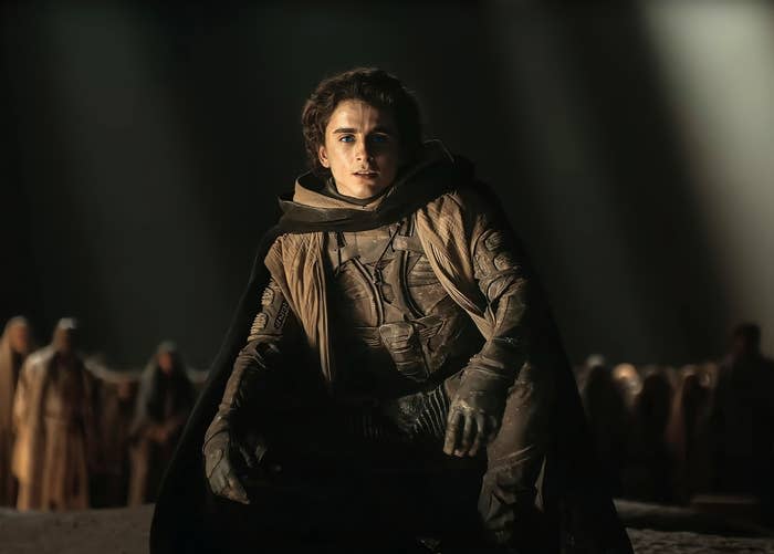 Paul Atreides in a textured coverall with a cloak stands center in a dramatic scene from the film "Dune"