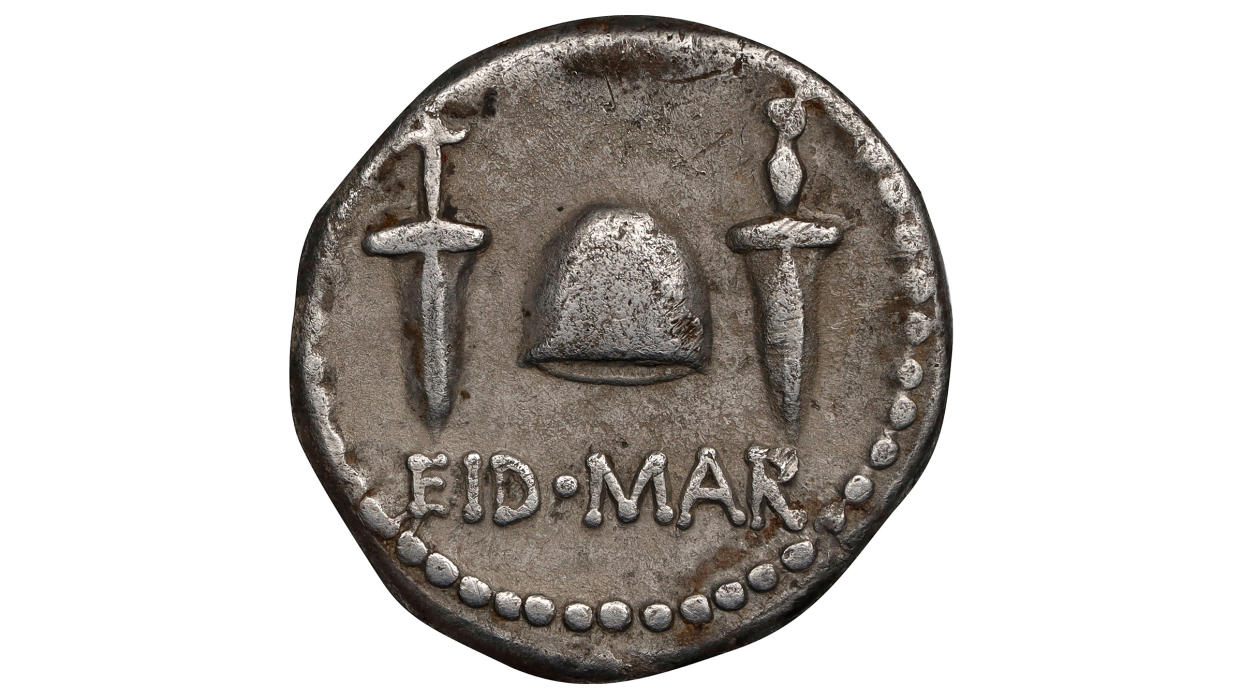  A silver coin against a white background. The coin has two daggers and a hat between them. It says EID MAR for the ides of march. 