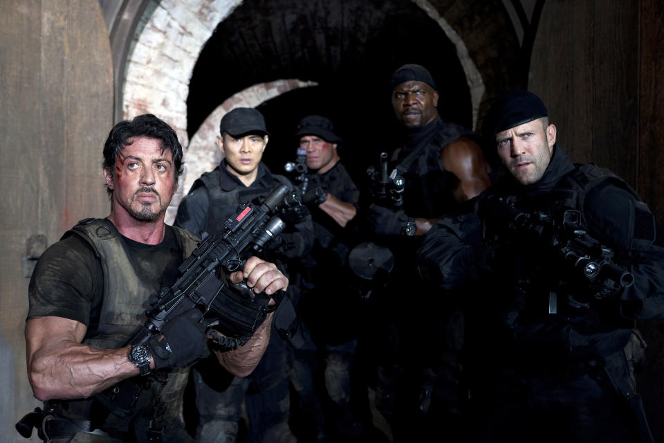 Sylvester Stallone, Jet Li, Randy Couture, Terry Crews, and Jason Statham stand armed in a hallway