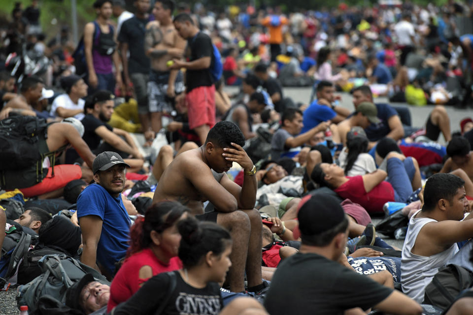 Scores of migrants, some looking exhausted, sit together on the ground.