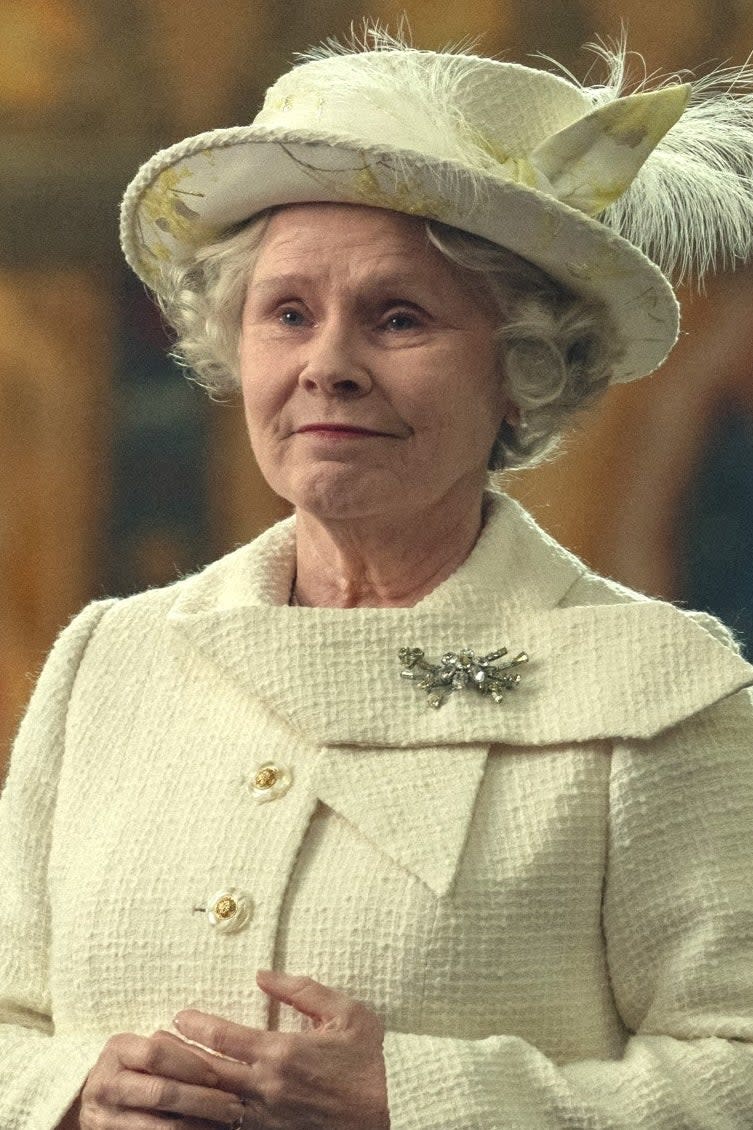 Imelda Staunton, dressed in regal attire with a hat featuring feathers, stands in front of an ornate, historic backdrop, evoking a scene from a period drama