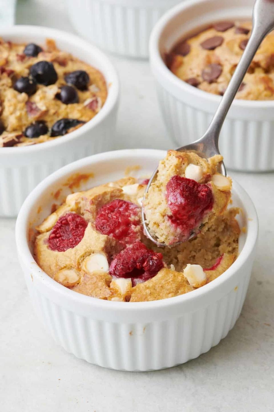 A spoon lifting a portion of baked oats with raspberries and white chocolate chips, served in a white ramekin, with three more similar dishes in the background