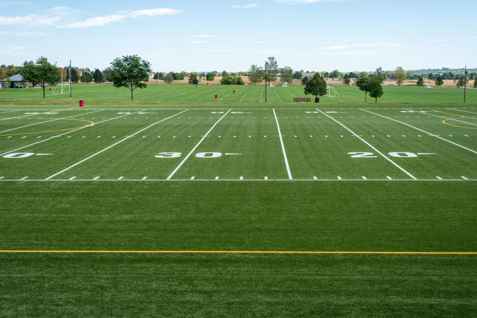 An empty, neatly maintained football field with visible yard markings, goal posts, and a few scattered trees in the background