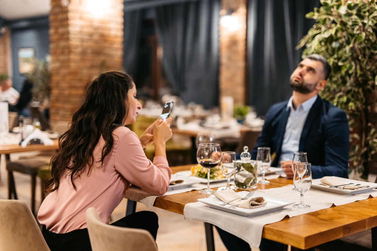 Mid adult woman using smart phone while her husband is frustrated at the restaurant.