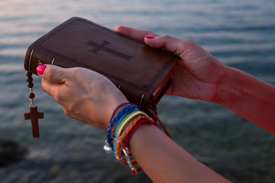 Hands holding a Bible with a cross and rosary, adorned with rainbow bracelets, against a background of water