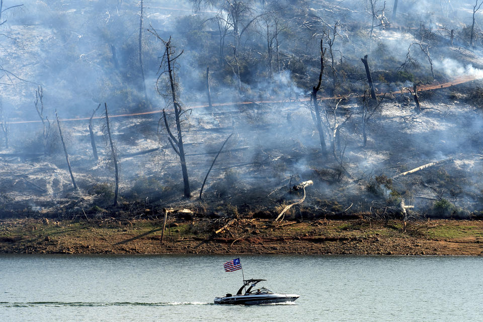 A boat crosses Lake Oroville with a smoldering hillside behind as the Thompson Fire burns in Oroville, Calif., on Wednesday, July 3, 2024. An extended heatwave blanketing Northern California has resulted in red flag fire warnings and power shutoffs. (AP Photo/Noah Berger)