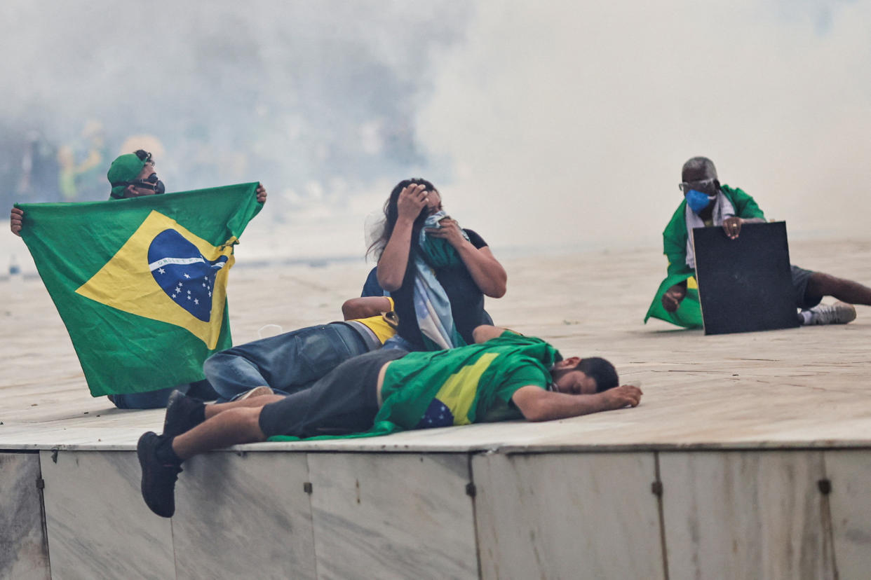 Supporters of Brazil's former President Jair Bolsonaro sit on the ground with clouds of tear gas in the background. One is prostrated on the cement.