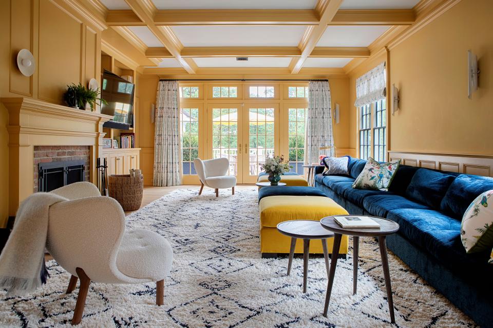 In this stylish living area, a TV was placed to the side of the fireplace in order to “make it a little bit out of the way and more gracious,” says Galli. Above the fireplace are sconces by Allied Maker. The indigo sofa, from Mitchell Gold + Bob Williams, is paired with two ottomans upholstered in Maharam fabric. The window treatments, in fabric by Eskayel, break up the walls, which were painted in a yellow hue by Farrow & Ball. The white chairs are by Matter, and the custom Moroccan rug is from Anou.