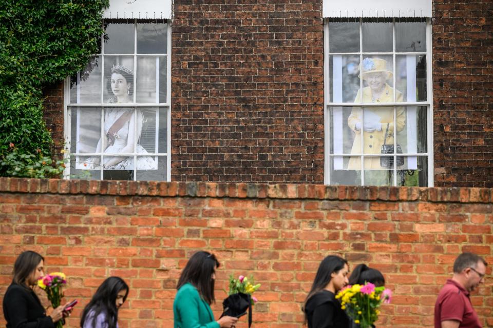 People walk with flowers in front of two windows at Windsor Castle that are decorated with pictures of the Queen.