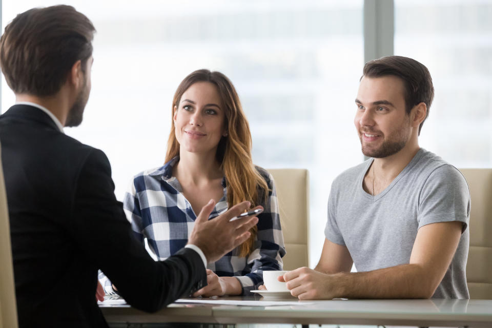 A couple smiles while sitting at a desk as a man in a suit provides them with information.