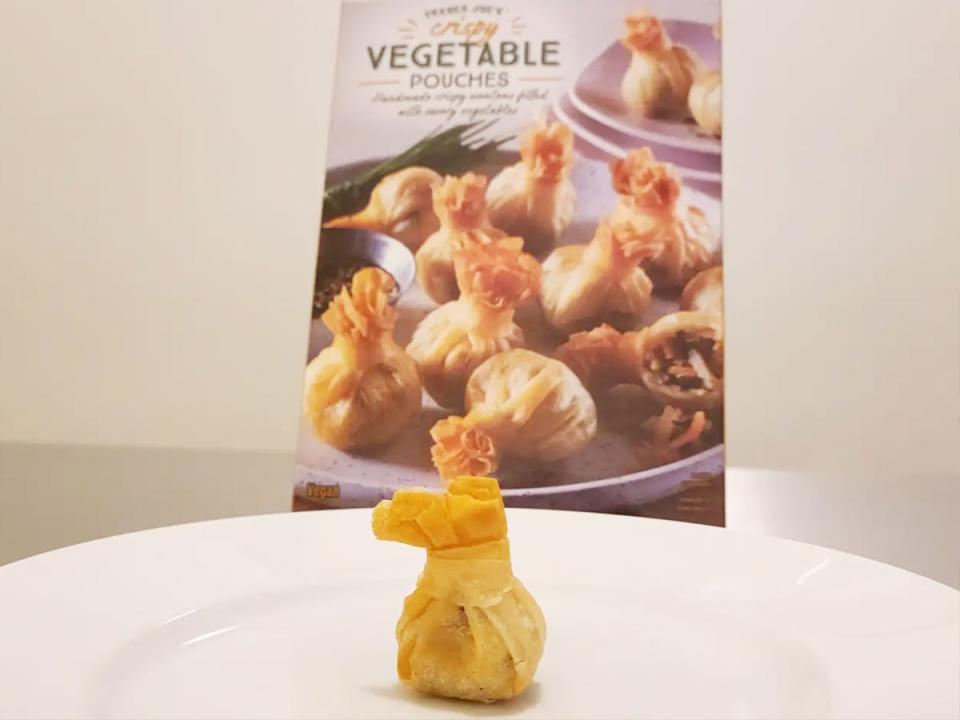 A golden-brown vegetable pouch with crispy ends sits on a white plate in front of a gray box