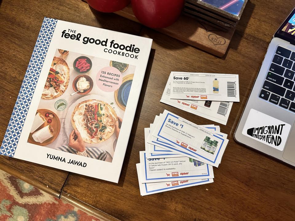 A cookbook titled "The Feel Good Foodie Cookbook" on a table with coupons and a laptop nearby
