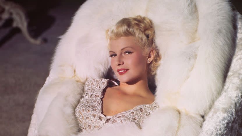 rita hayworth sits in a white chair wearing a large white fur coat and white and silver top, she smiles and eyes the camera