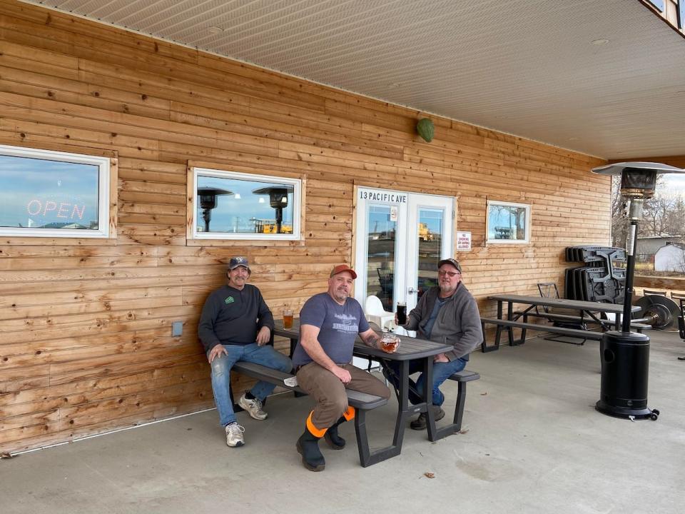 Rafter R Brewing Company owner and brewer Ryan Moncrieff, centre, regular customer Jim Roseveare, left, and regular customer Bob Hamenjoy enjoy their brews as they bask on the Maple Creek business's outside patio Tuesday afternoon.  (Rafter R Brewing Company  - image credit)