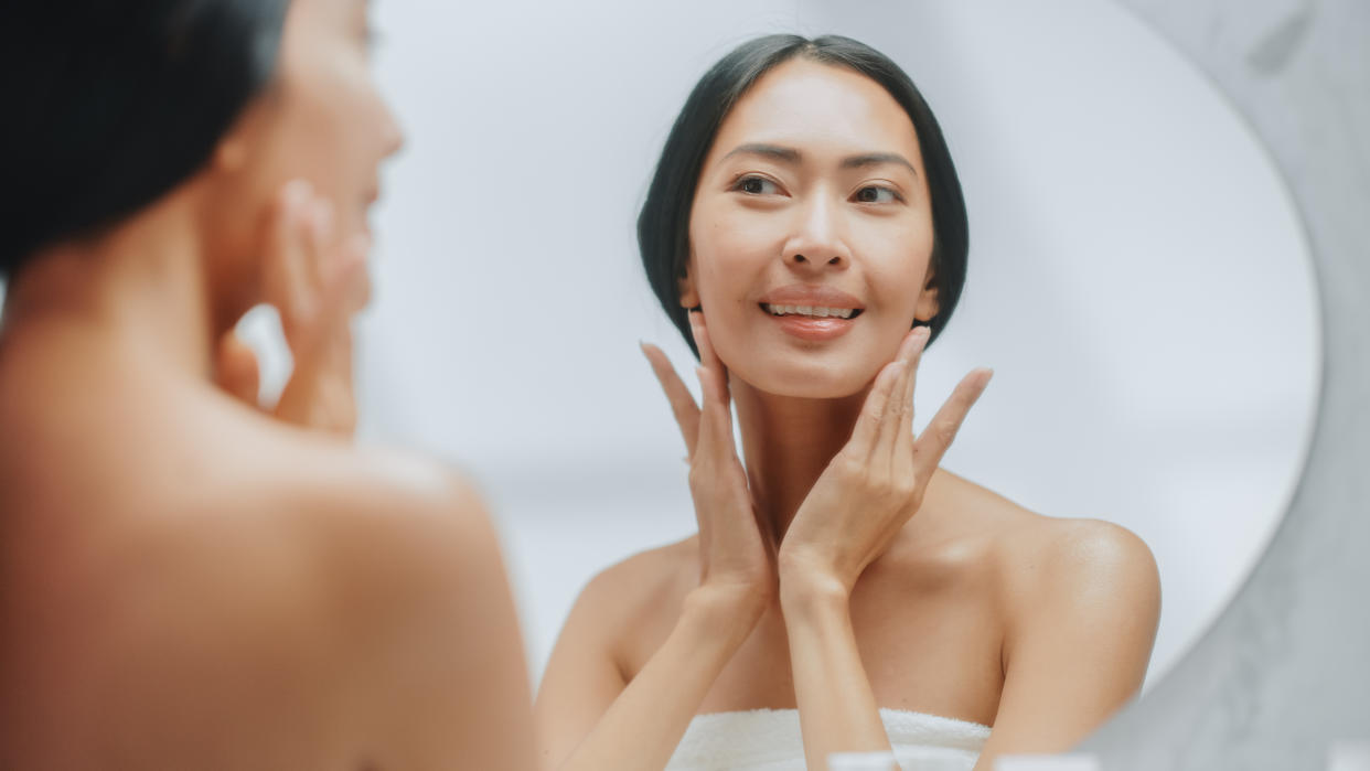 A photo of a woman applying face cream and looking at the bathroom mirror. 