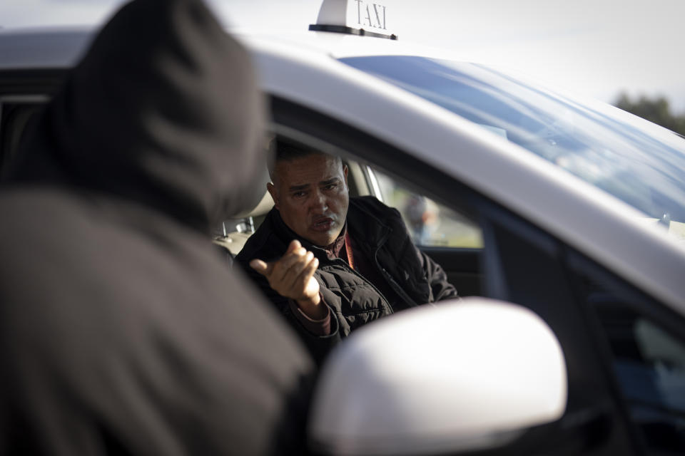 A taxi driver offers rides to migrants as they arrive at a bus stop after leaving a processing facility, Friday, Feb. 23, 2024, in San Diego. Hundreds of migrants were dropped off Friday at a sidewalk bus stop amid office parks in San Diego with notices to appear in immigration court after local government funding for a reception center ran out of money sooner than expected. (AP Photo/Gregory Bull)