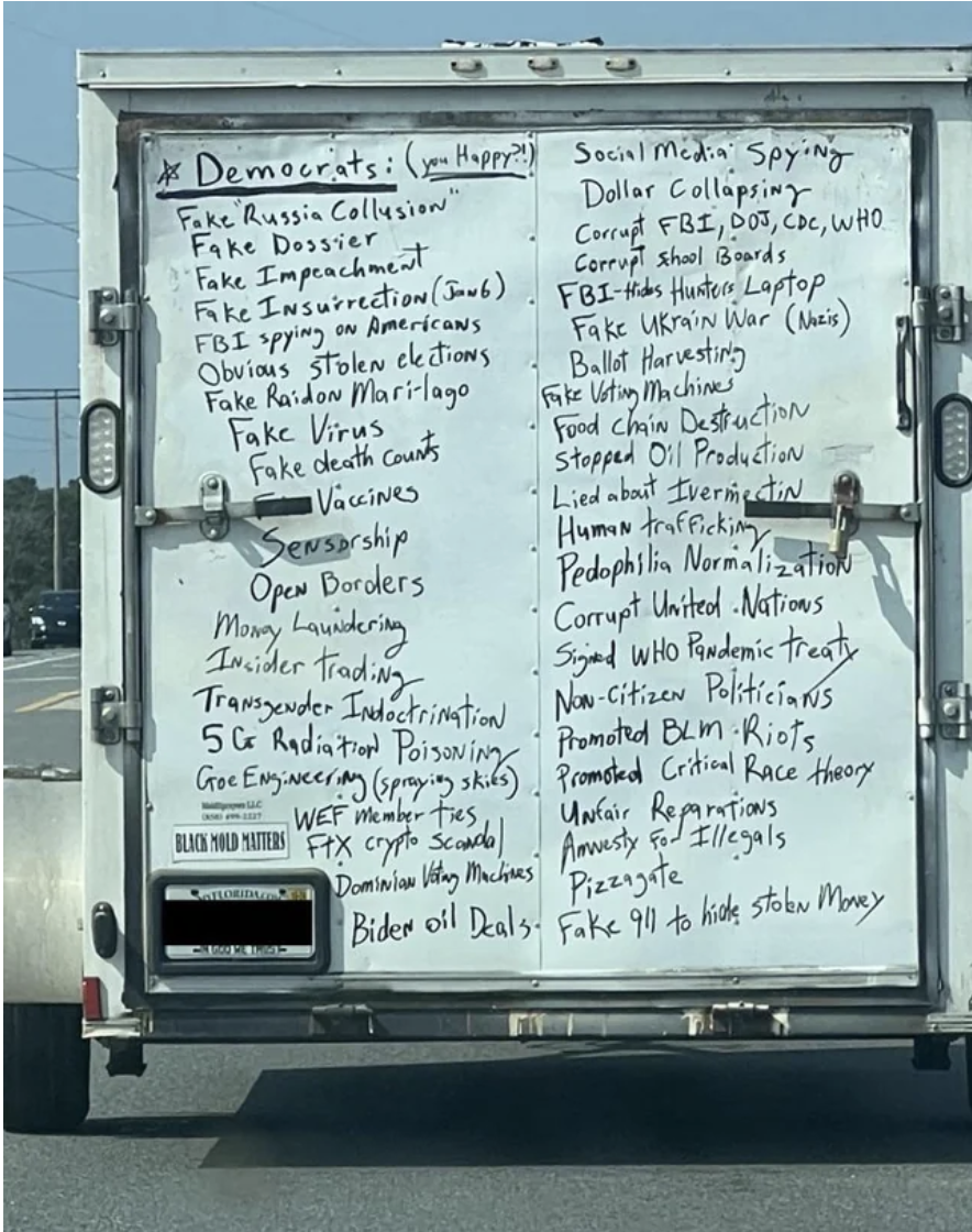 The back of a truck on the highway with a long handwritten list under the heading "Democrats," including fake dossier, fake virus, fake impeachment, Dominion voting machines, transgender indoctrination, unfair reparations, and pizzagate