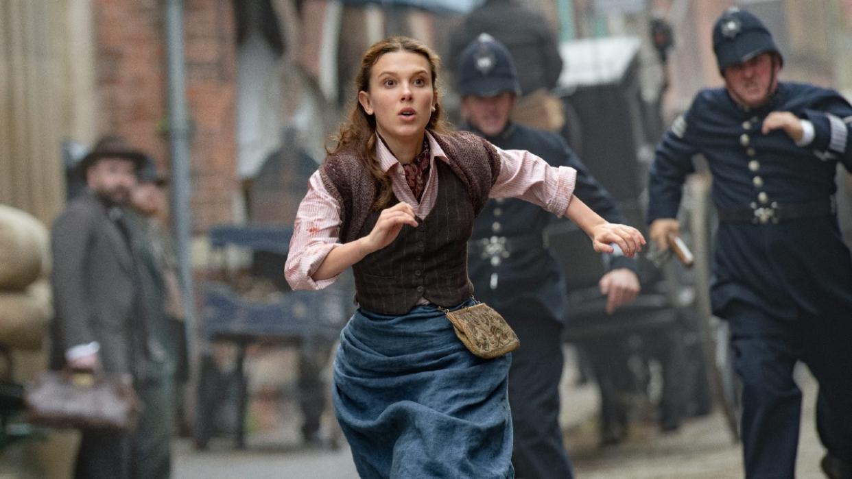  A press image of Millie Bobby Brown as Enola being chased by cops down a street in Enola Holmes 2. 