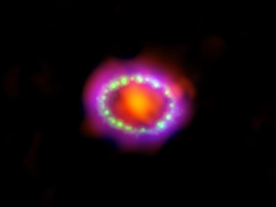 supernova with dark orange center surrounded by pearls of green and blue light inside a purple cloud against the black background of space