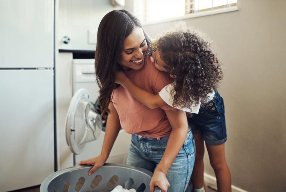 A child leans on an adult's shoulder while they do laundry.