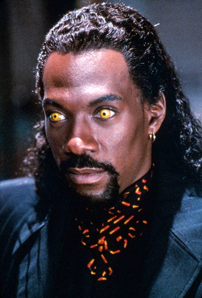 Eddie Murphy in character with glowing yellow eyes and dressed in a dark suit, accessorized with a patterned scarf