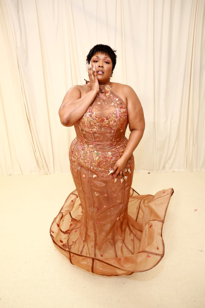 Lizzo in a transparent floral embroidered dress with a halter neckline, posing with her hand on her face against a neutral background