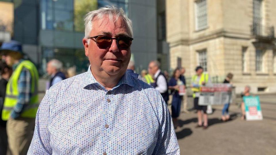 Peter Richardson looks into the camera wearing sunglasses in front of campaigners