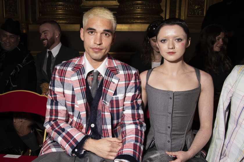 A man with platinum hair in a plaid coat sitting next to a woman with short hair in a grey sleeveless dress