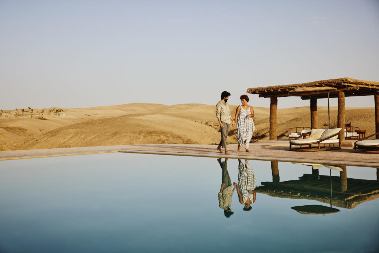 2023 Travel Noire Awards: Favorite Unique Stay. Wide shot of couple walking around pool at Moroccan desert camp at sunrise while on vacation