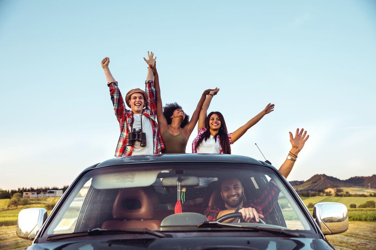 Happy four young adults traveling in a car, three are hanging out of the sunroof while one male is at the driver's seat, arms are in a 'V', with a idyllic rural background