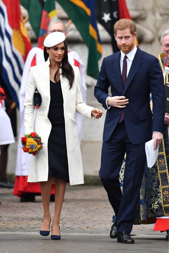 Meghan in a white coat and beret and navy knee-length dress and Harry in a Navy suit in front of colorful flags.