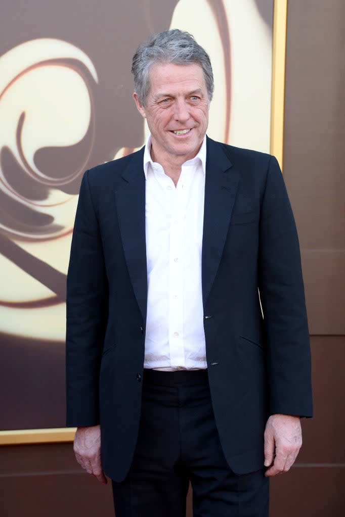 Hugh Grant is smiling while posing on the red carpet, wearing a suit jacket over a dress shirt