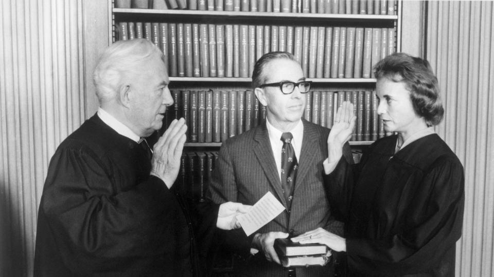 warren burger and sandra day o'connor stand facing each other each with their right hand raised, a man stands in the middle holding a stack of books that o'connor rests her left hand on