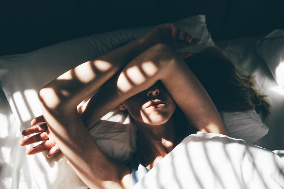 A woman laying in bed with her arms covering her eyes, as the light shines through blinds in her bedroom. (Photo via Getty Images)