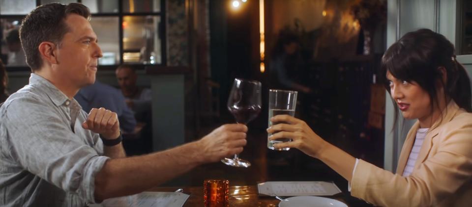 A man and a woman with dark hair sit across a table at a restaurant. He has red wine. She has a glass of water. They cheers.