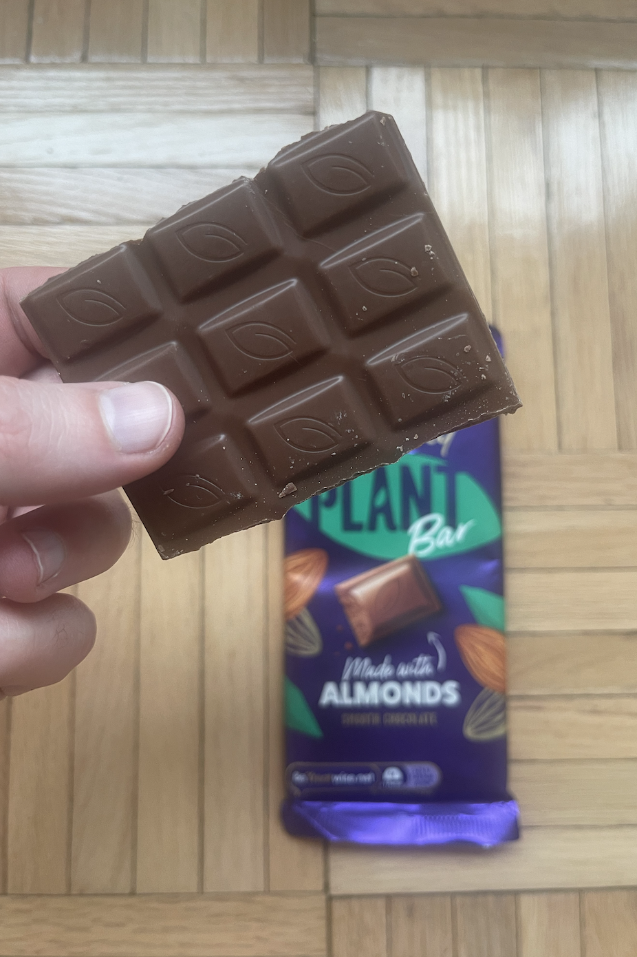 Hand holding a partially unwrapped chocolate almond bar over a wooden floor with the wrapper visible