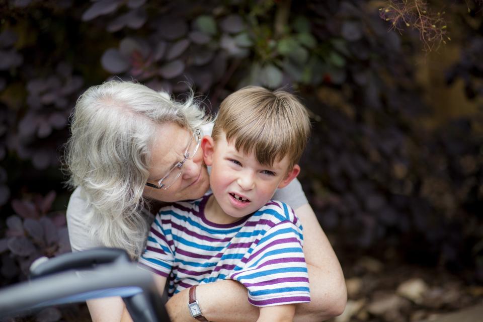 An elderly woman with gray hair embraces a young boy wearing a striped shirt in a garden