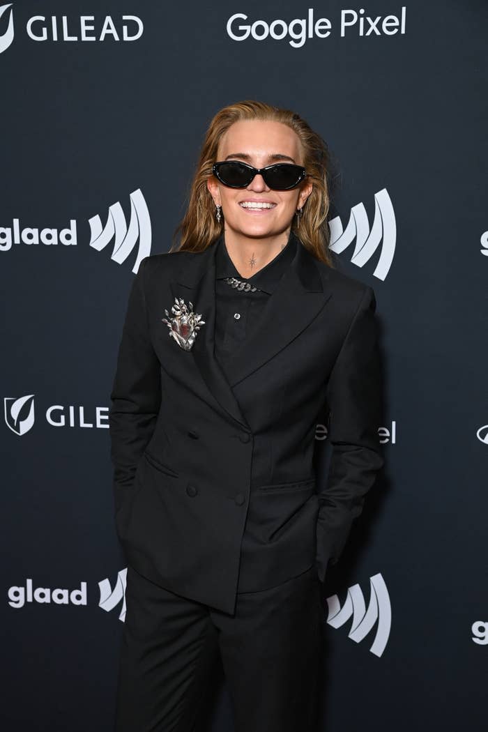 Person on a red carpet in a black suit with a brooch and sunglasses at the GLAAD event. Logos for "GLAAD," "Google Pixel," and "Gilead" are in the background