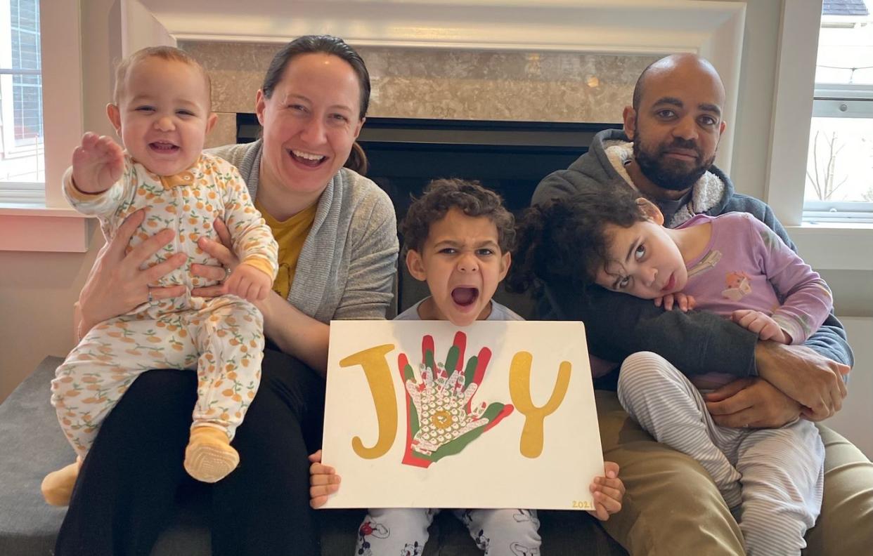 Mom and dad with three young children smiling, while one holds a sign that says 