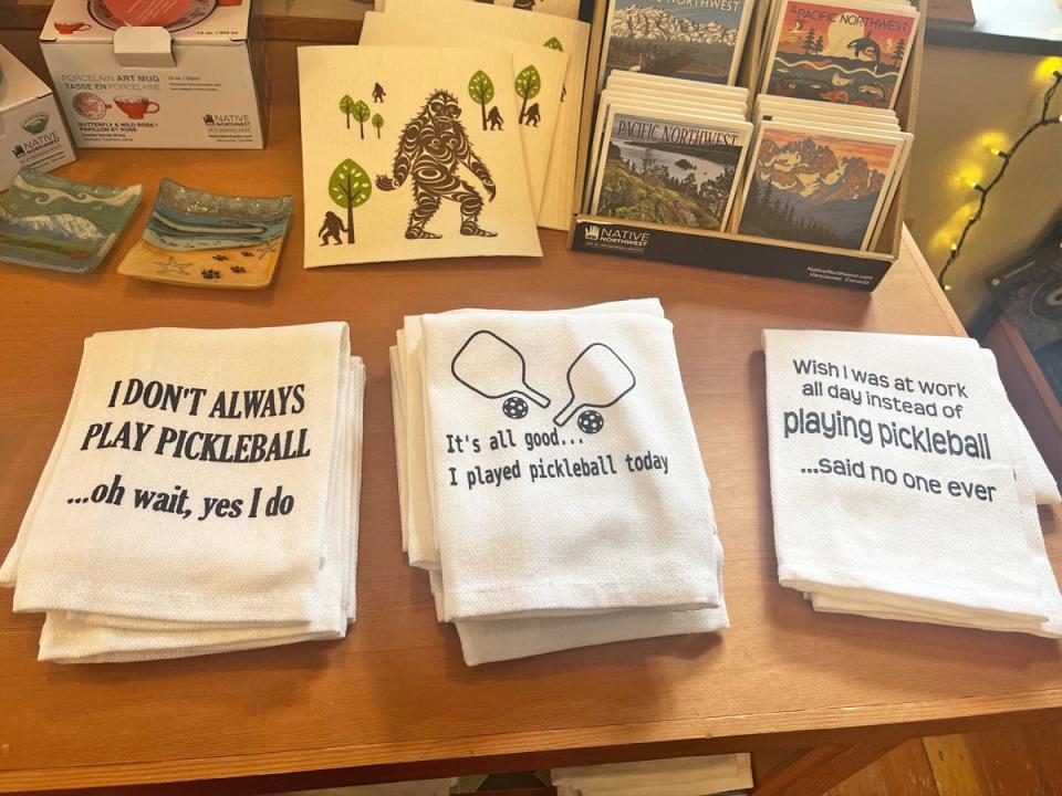 Three white clothes on a wooden table with pickleball-themed sayings and illustrations of pickleball paddles