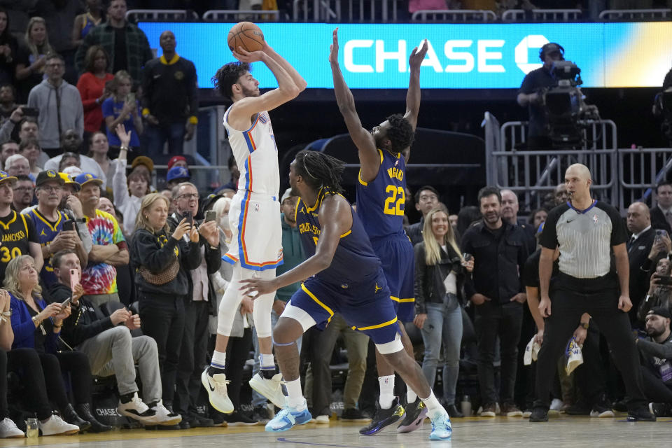 Oklahoma City Thunder forward Chet Holmgren, left, makes a 3-point basket while being defended by Golden State Warriors forward Andrew Wiggins (22) and forward Kevon Looney during the second half of an NBA basketball game in San Francisco, Saturday, Nov. 18, 2023. The Thunder won 130-123 in overtime. (AP Photo/Jeff Chiu)