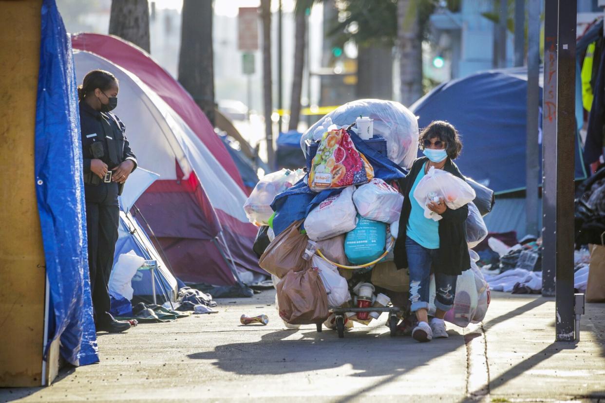 A uniformed officer watches from a row of tents as a woman pulls a cart with a pile of bags reaching above her head.