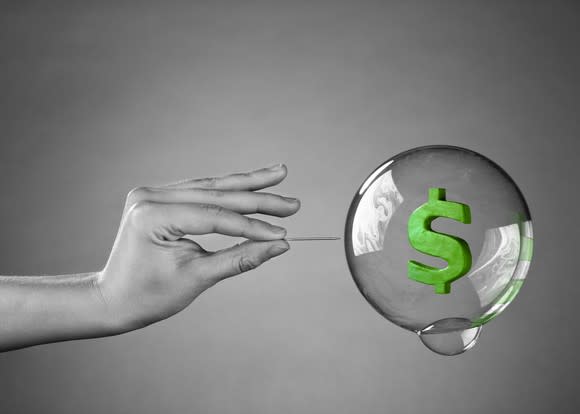 green dollar sign in a clear bubble, with a hand holding a pin about to burst it