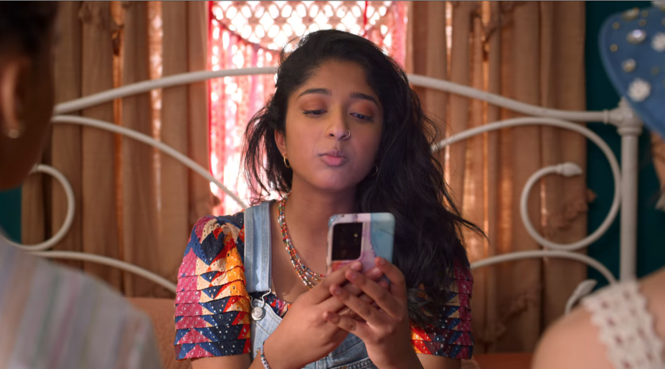 Devi from "Never Have I Ever" on Netflix sits on her bed, looks at her phone as if texting or on an app.