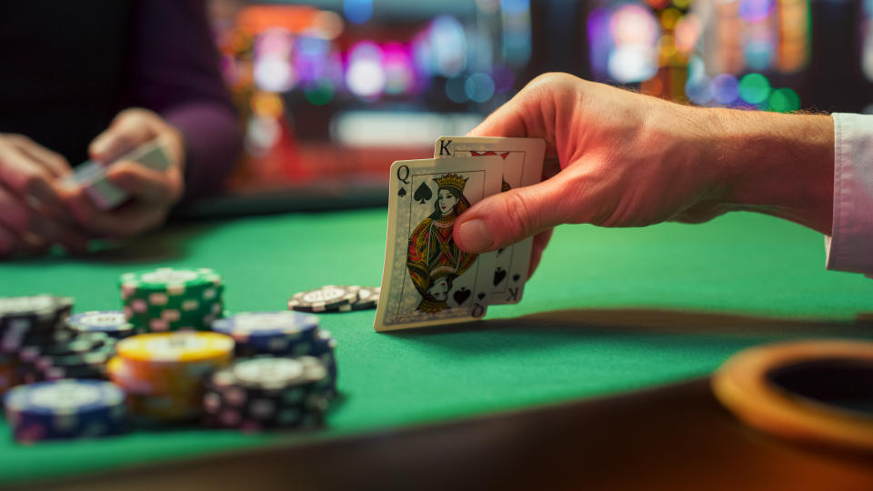 A person holds two playing cards, the Queen of Spades and King of Clubs, at a poker table with poker chips stacked in front of them