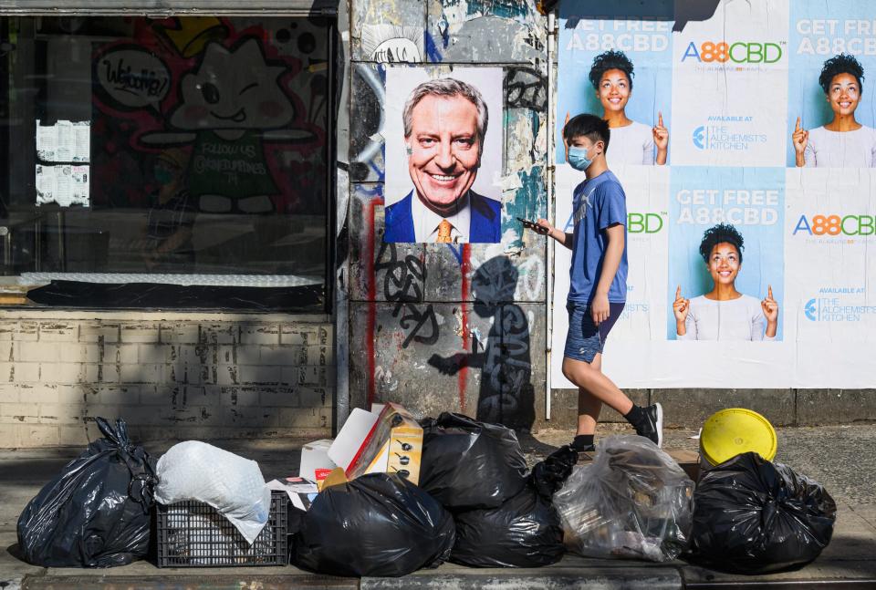 A masked man is seen walking by a pile of trash and a poster of the New York mayor.