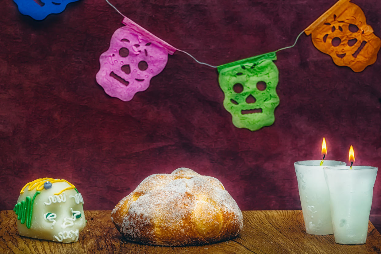 Pan de muerto en fondo adornado con veladoras y calabera de azucar, papel picado en el fondo