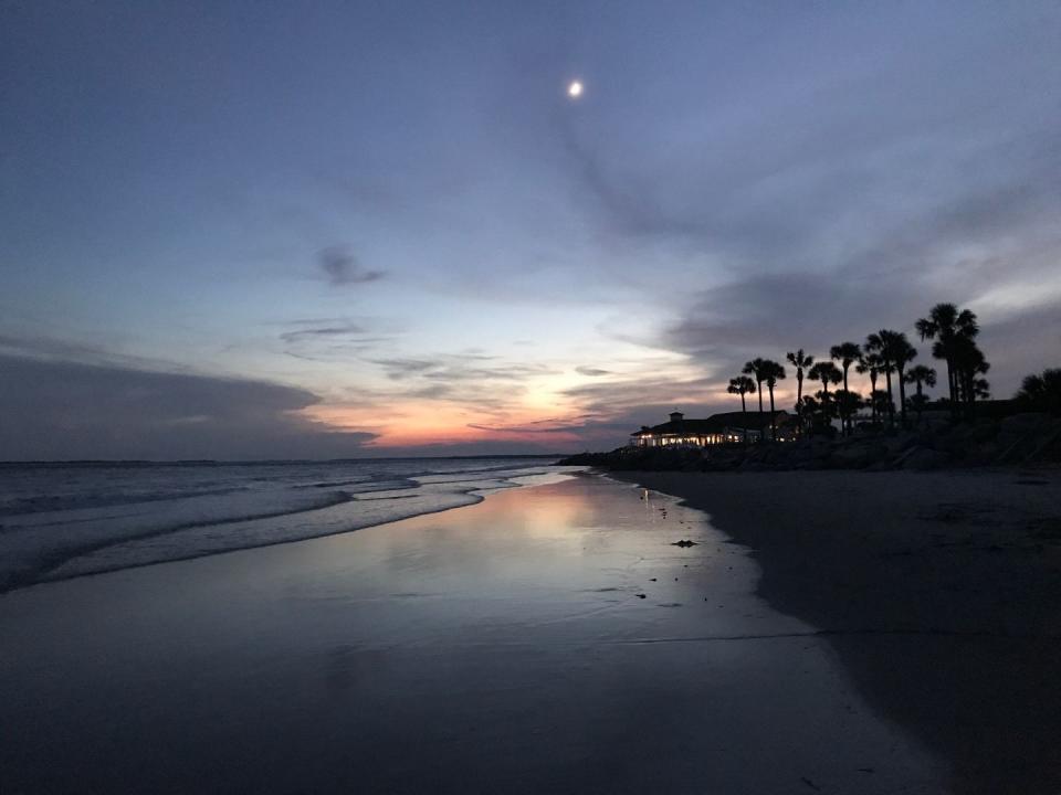 scenic view of beach during sunset with moon, outline of palm trees and lighted structure in the distance