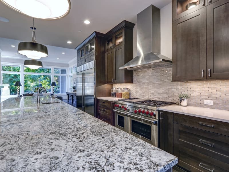 Modern kitchen with brown kitchen cabinets, oversized kitchen island, granite countertops, stainless steel hood over six burner Range and beige backsplash. Northwest, USA